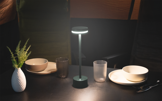 Kleine Tischszene mit zwei gedeckten Plätzen, jeweils Teller und Schale, zwei Gläser, runde Tischlampe in der Mitte und Vase mit grünen Zweigen links