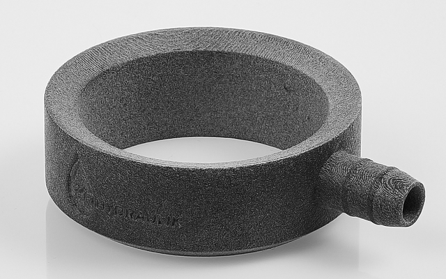 Objeto en forma de anillo gris oscuro con un corto cilindro lateral, sobre fondo blanco con sombra tenue