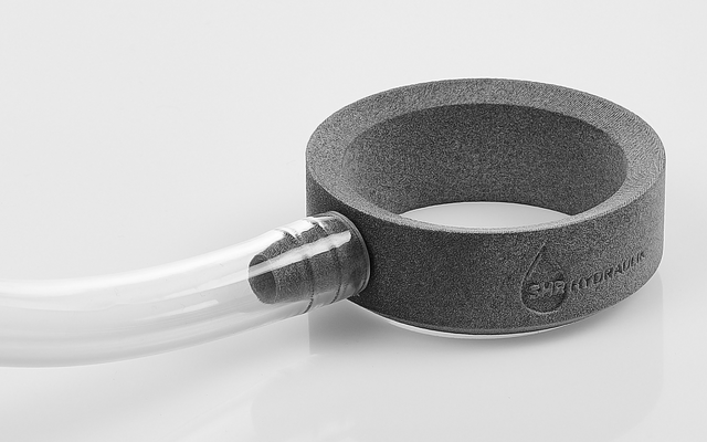 Anillo gris de espuma con una manguera transparente conectada mediante un conector metálico sobre fondo blanco