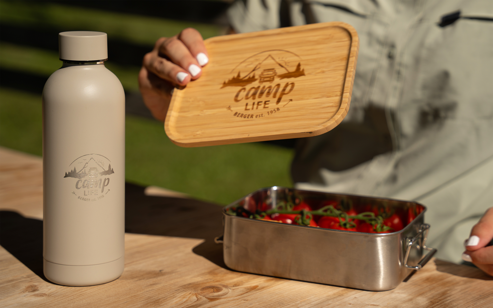 Main aux ongles vernis blanc tient un couvercle en bois avec l'inscription "Camp Life" au‑dessus d'une boîte en acier inoxydable contenant des fraises; une bouteille beige avec le même logo est posée sur une table en bois