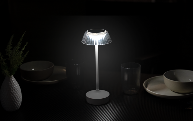 Kleine Tischlampe mit geripptem, transluzentem Schirm und rundem Sockel auf dunkler, strukturierter Oberfläche; umgeben von Tellern, Schalen, Trinkgläsern und einer weißen Vase mit Zweigen