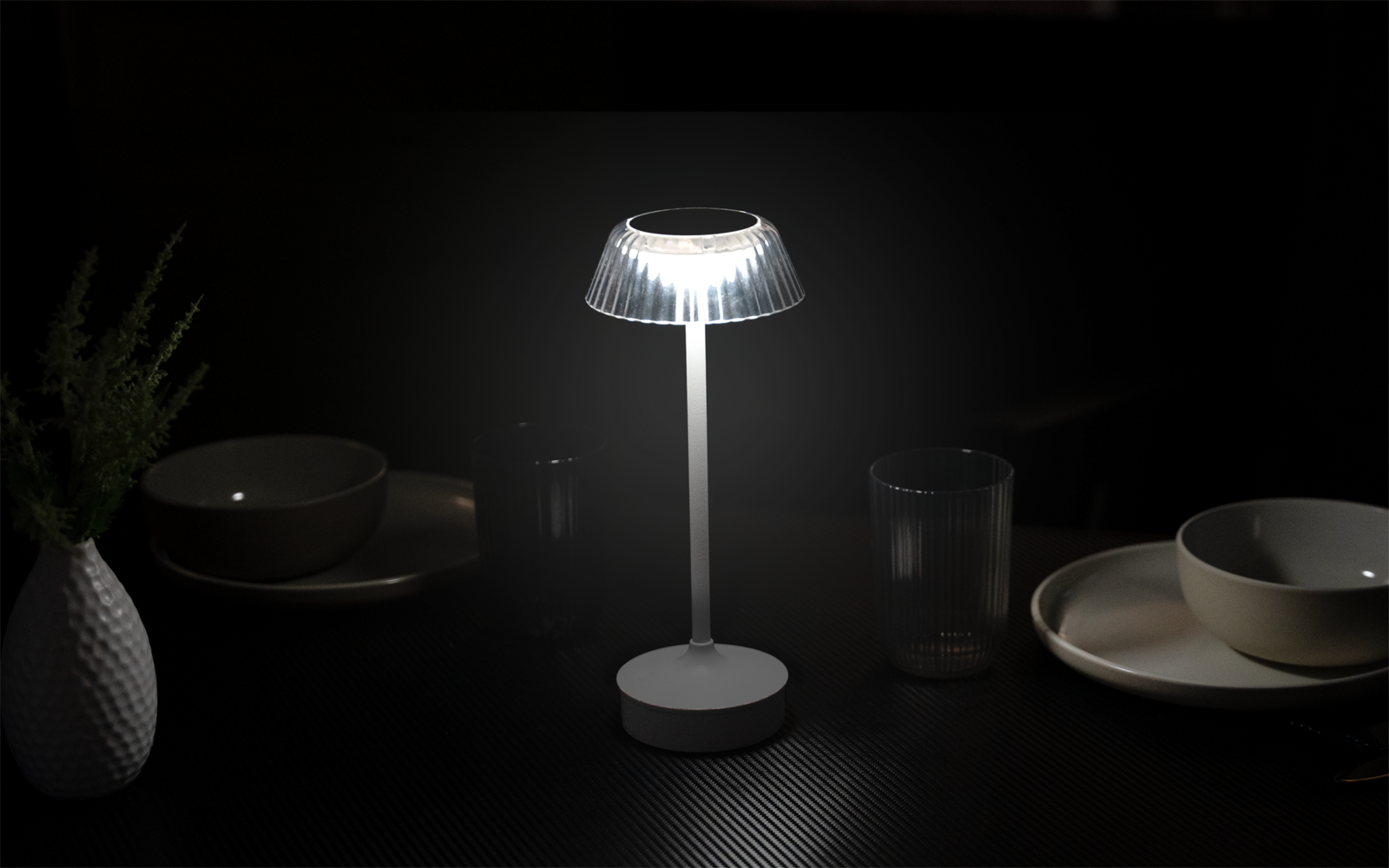 Kleine Tischlampe mit geripptem, transluzentem Schirm und rundem Sockel auf dunkler, strukturierter Oberfläche; umgeben von Tellern, Schalen, Trinkgläsern und einer weißen Vase mit Zweigen