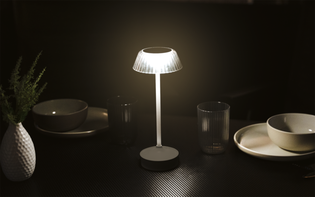 Weiße Tischlampe mit beleuchtetem, transparentem Schirm auf dunklem Tisch, umgeben von Tellern, Schalen, zwei Gläsern und einer kleinen Vase mit Zweigen