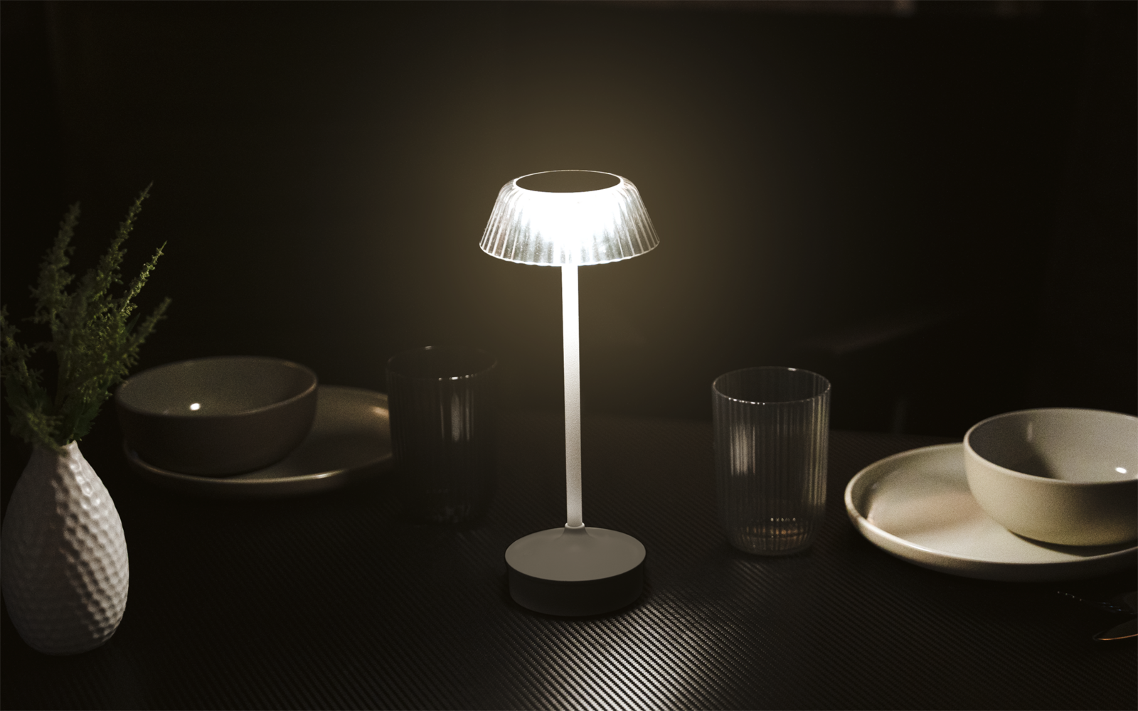 Weiße Tischlampe mit beleuchtetem, transparentem Schirm auf dunklem Tisch, umgeben von Tellern, Schalen, zwei Gläsern und einer kleinen Vase mit Zweigen