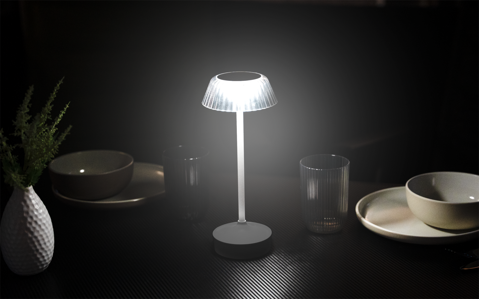 Kleine leuchtende Tischlampe mit geriffeltem, durchscheinendem Lampenschirm auf rundem Sockel auf dunklem Tisch; umgeben von Schalen, Tellern, einem Glas und einer weißen Vase mit grünem Zweig