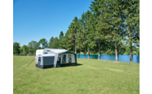 Weißer Wohnwagen mit angebautem Vorzelt auf grüner Wiese neben einem See, hohe Nadelbäume im Hintergrund und blauer Himmel
