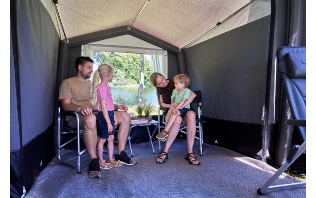Intérieur d'une annexe de camping avec quatre personnes : deux hommes adultes assis sur des chaises pliantes, une fillette debout, un tout-petit sur les genoux d'un homme ; petite table avec une plante au centre ; arrière ouvert donnant sur des arbres et de l'eau