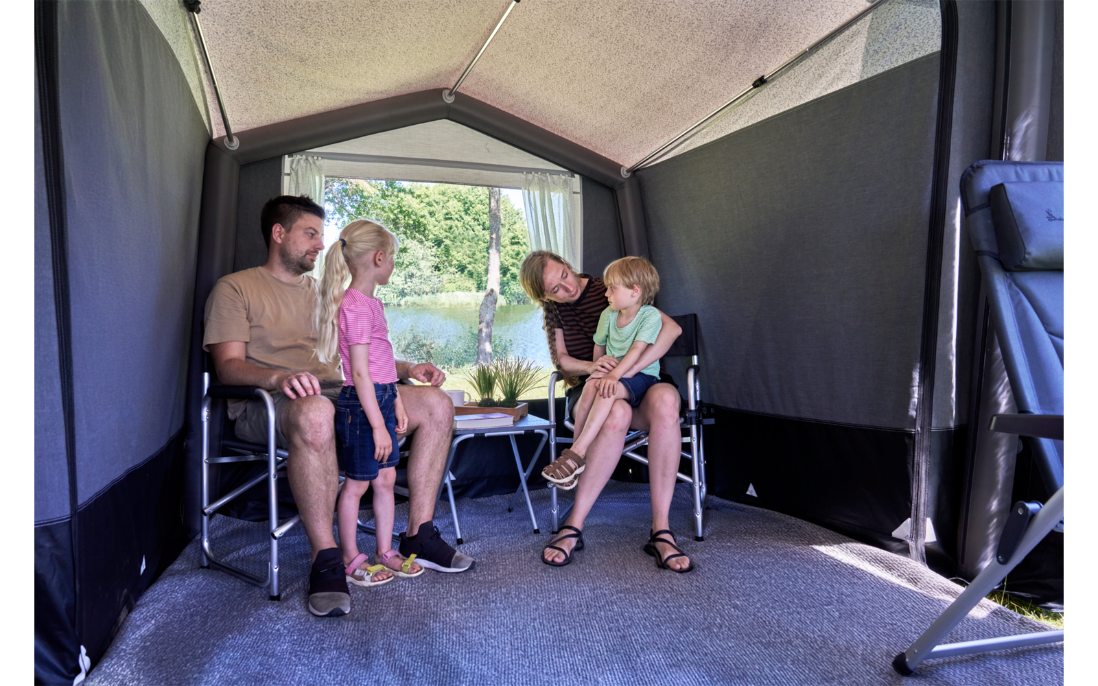 Intérieur d'une annexe de camping avec quatre personnes : deux hommes adultes assis sur des chaises pliantes, une fillette debout, un tout-petit sur les genoux d'un homme ; petite table avec une plante au centre ; arrière ouvert donnant sur des arbres et de l'eau