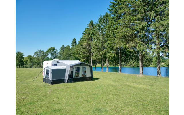 Caravane blanche avec auvent fixé sur une pelouse près d'un lac, grands conifères en arrière‑plan et ciel bleu