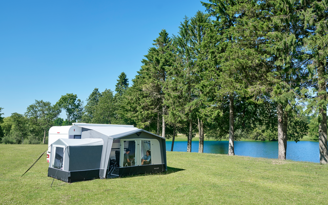 Weißer Wohnwagen mit angebautem Vorzelt auf grüner Wiese neben einem See, hohe Nadelbäume im Hintergrund und blauer Himmel