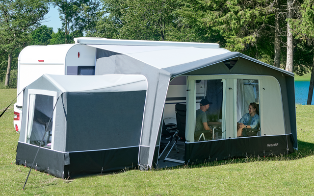 Weißer Wohnwagen mit angebautem Vorzelt auf grüner Wiese neben einem See, hohe Nadelbäume im Hintergrund und blauer Himmel