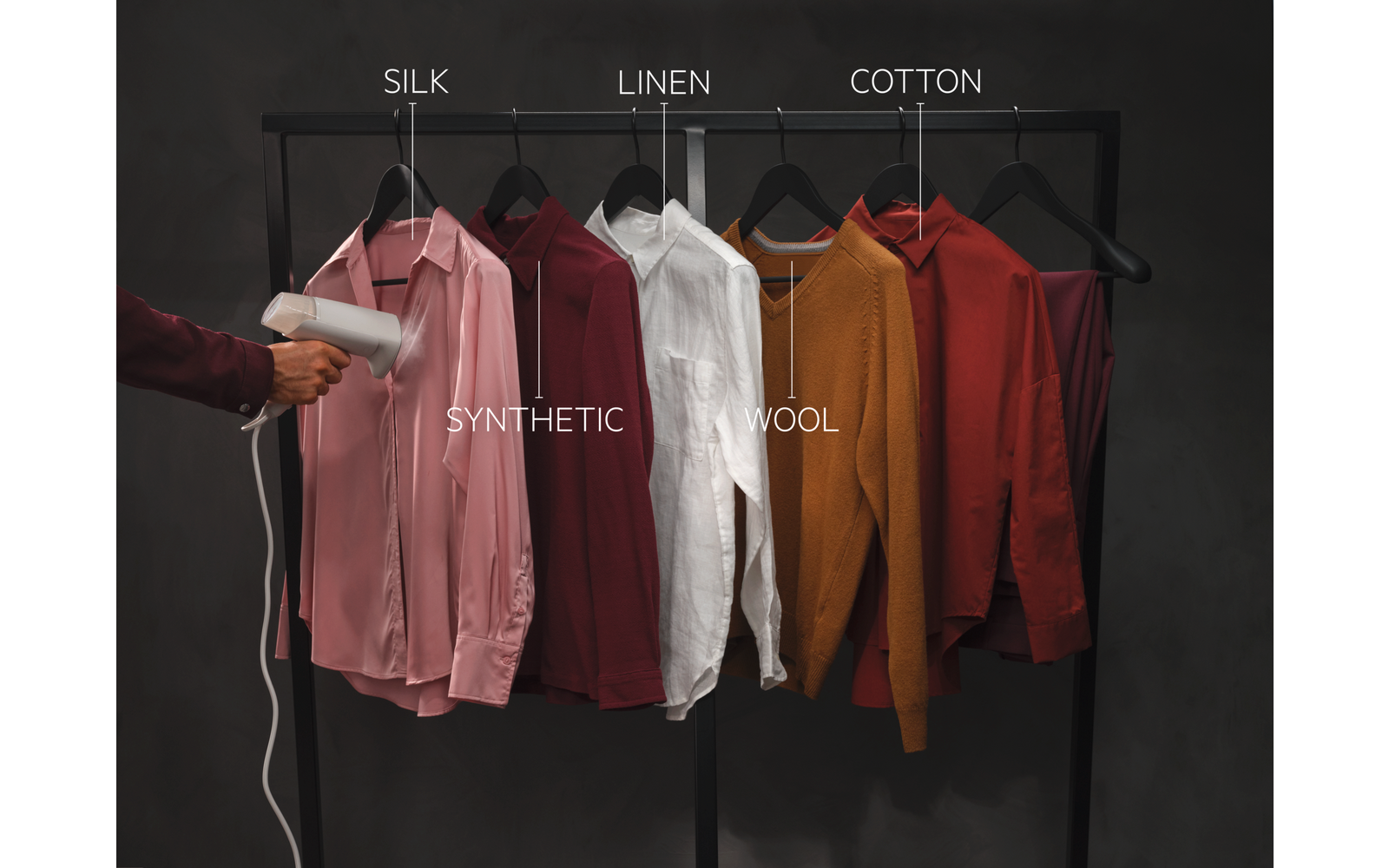 Kledingrek met hangende bovenkleding in verschillende kleuren, zichtbare teksten 'SILK', 'SYNTHETIC', 'LINEN', 'WOOL', 'COTTON', links een hand met een handstomer