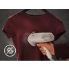 Hand houdt een wit handstoomapparaat tegen een bordeauxrood T‑shirt aan een hanger; stoom zichtbaar; linksonder een rond pictogram met doorgestreepte kiemen