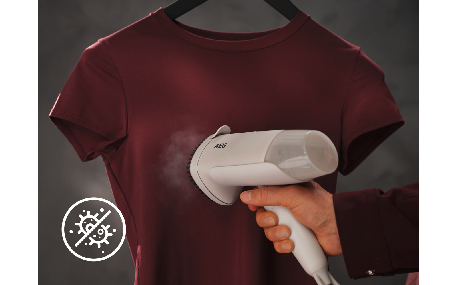 Hand houdt een wit handstoomapparaat tegen een bordeauxrood T‑shirt aan een hanger; stoom zichtbaar; linksonder een rond pictogram met doorgestreepte kiemen