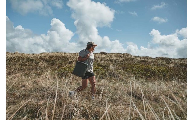 Persona caminando por dunas con hierba alta, lleva gorra, camiseta, pantalones cortos y bolso de hombro, cielo nublado