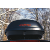 Box da tetto nero con logo rosso "KAMEI" montato su un veicolo; sullo sfondo rami di alberi innevati e cielo
