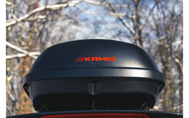 Box da tetto nero con logo rosso "KAMEI" montato su un veicolo; sullo sfondo rami di alberi innevati e cielo