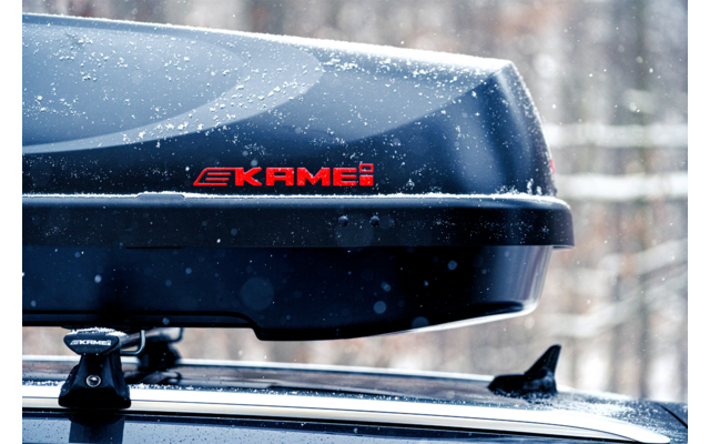 Box da tetto nero con logo rosso 'KAMEI' montato sul tetto dell'auto, coperto di neve, bosco sfocato sullo sfondo