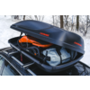 Box da tetto nero aperto con logo KAMEI sul tetto di un'auto; all'interno un casco arancione e una borsa grigio chiaro; bosco innevato sullo sfondo