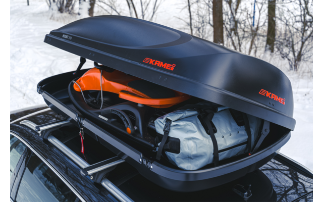 Box da tetto nero aperto con logo KAMEI sul tetto di un'auto; all'interno un casco arancione e una borsa grigio chiaro; bosco innevato sullo sfondo