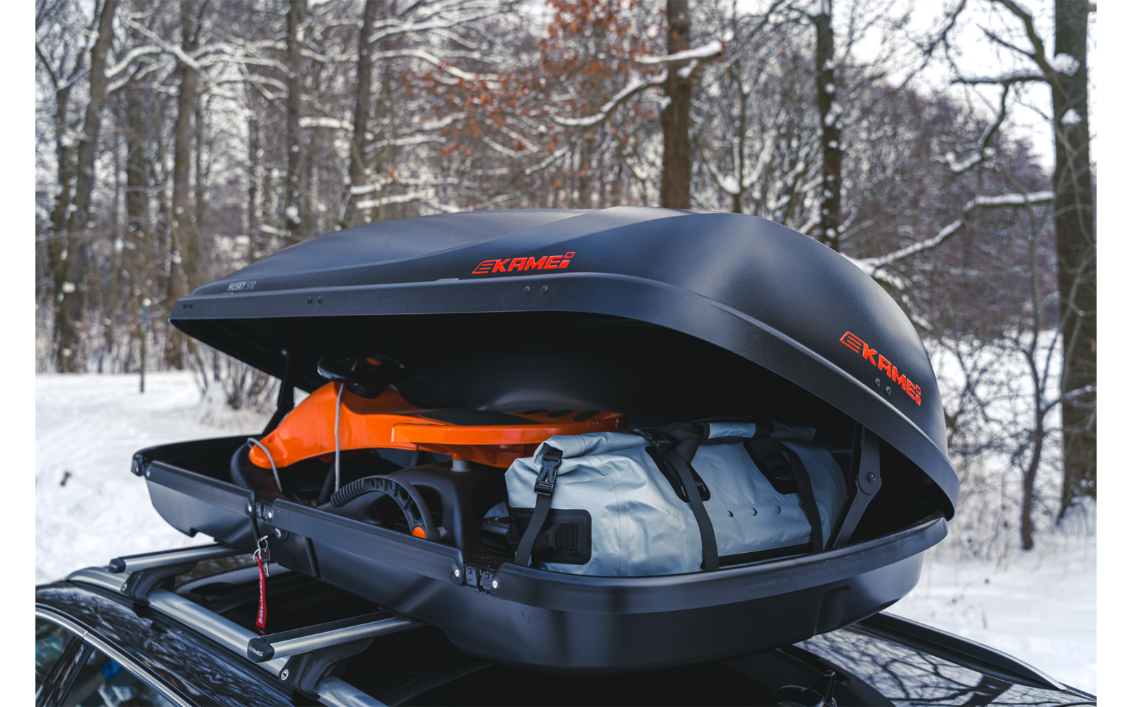 Box da tetto nero aperto sul tetto di un'auto con una borsa arancione e una valigia grigia, foresta innevata sullo sfondo