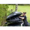 Persona apre il box da tetto di un'auto e mette la mano nel box con zaini; prato e alberi sullo sfondo