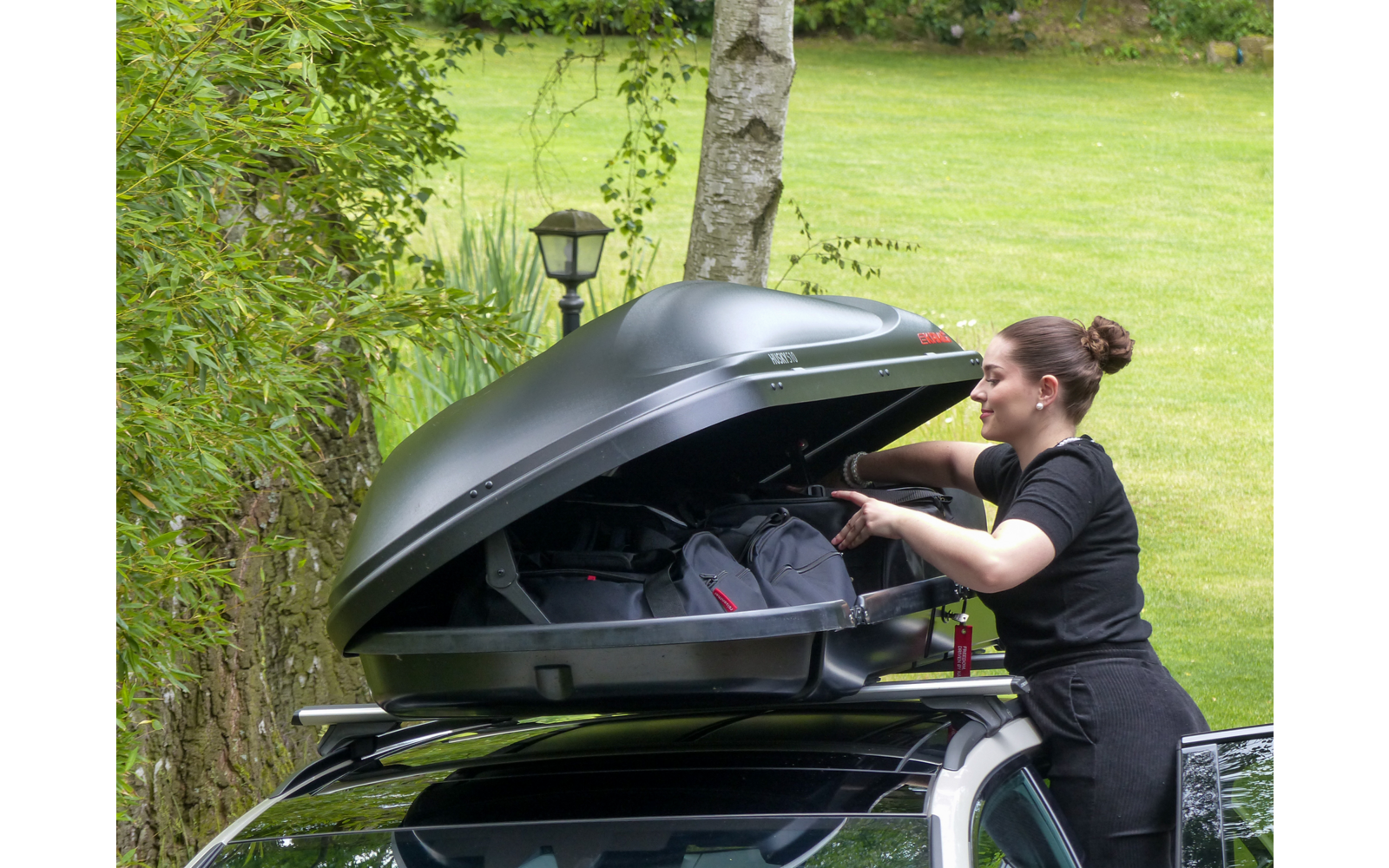 Persona apre il box da tetto di un'auto e mette la mano nel box con zaini; prato e alberi sullo sfondo