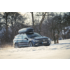 Station wagon scura con box sul tetto su una strada innevata, alberi spogli sullo sfondo