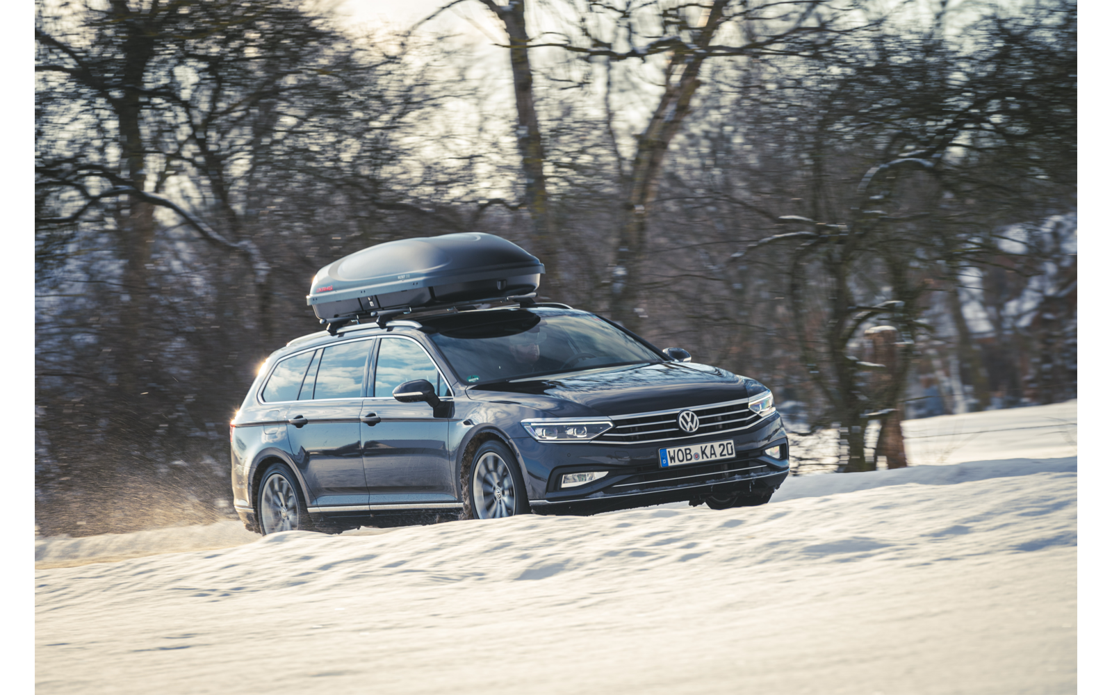 Station wagon scura con box sul tetto su una strada innevata, alberi spogli sullo sfondo