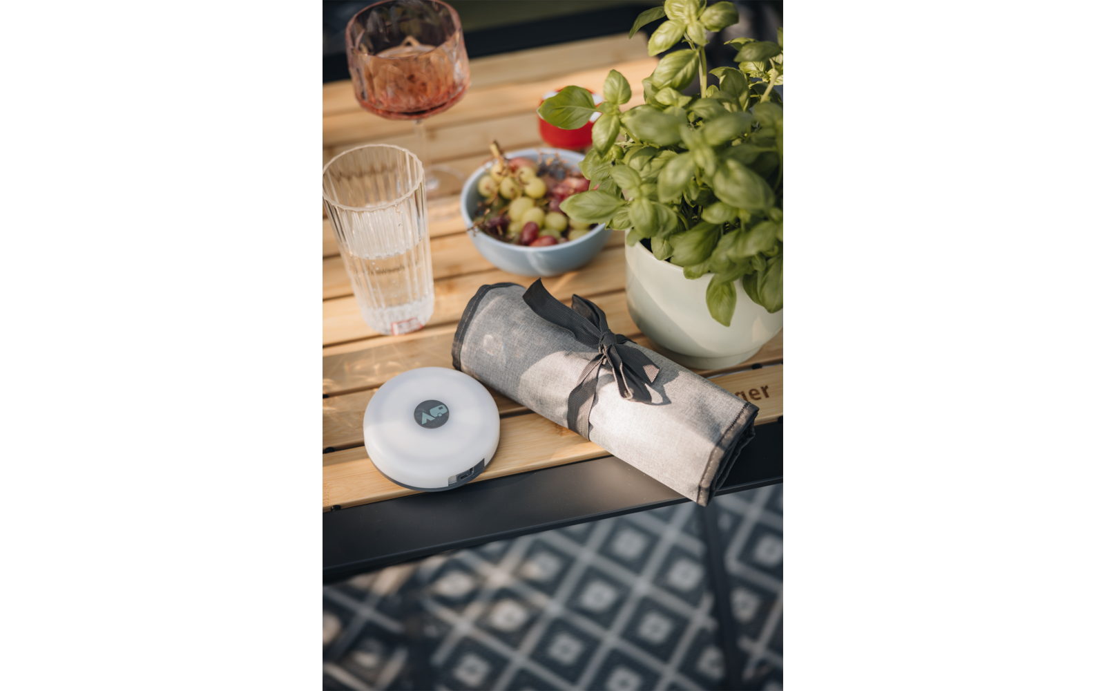 Holztisch mit Glas Wasser, Glas mit Getränk, Teller mit Oliven, Serviette mit Besteck und Pflanzen im Hintergrund
