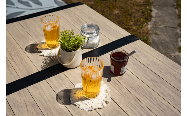 Holztisch im Freien, zwei gelbe Gläser mit Eis auf weißen gehäkelten Untersetzern; kleiner weißer Blumentopf mit grüner Pflanze; kleines Glas mit Deckel; Glas mit roter Marmelade und Löffel; Rasen und Steinweg im Hintergrund