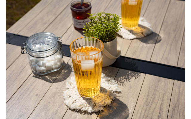 Zwei Gläser mit hellem Getränk und Eiswürfeln auf Makramee-Untersetzern auf einem Holztisch; daneben ein Glas mit Zuckerwürfeln, ein Glas mit dunklem Inhalt und eine kleine grüne Topfpflanze