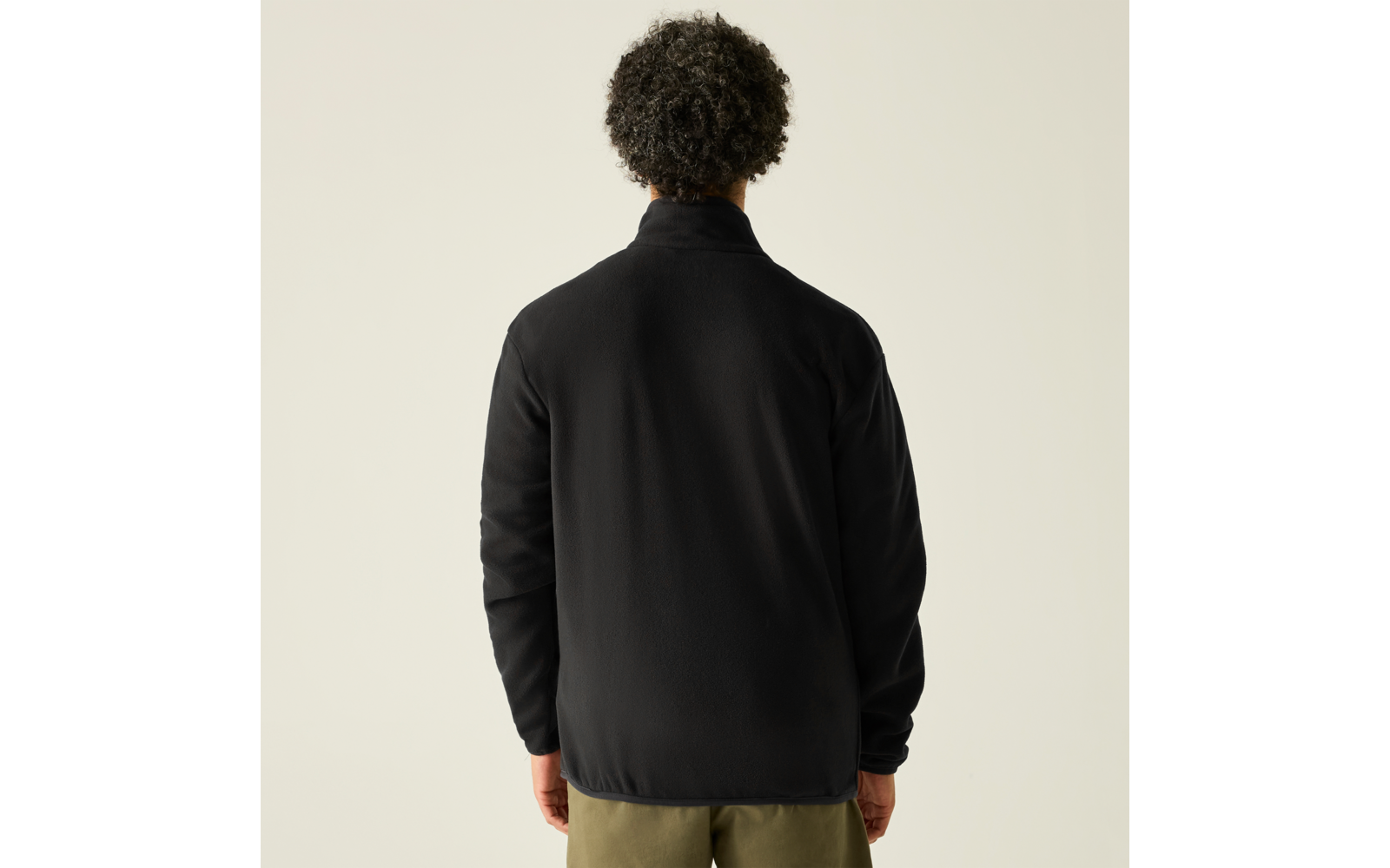 Person von hinten, trägt schwarze Jacke und olivgrüne Hose, lockige Haare, heller Hintergrund