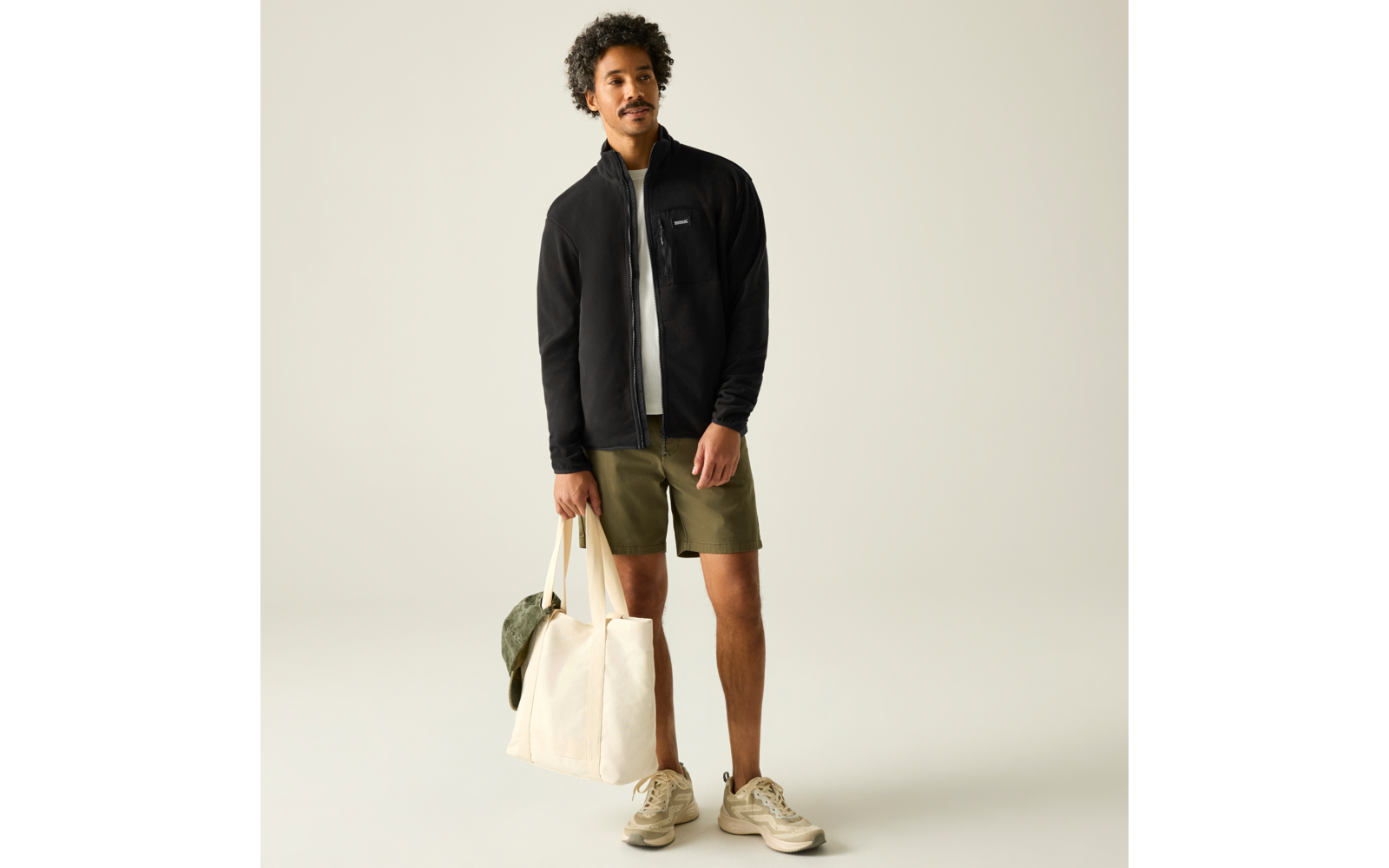 Person steht in schwarzer Jacke mit Reißverschluss, weißem T-Shirt, olivgrünen Shorts und beigen Sneakern, hält eine große beige Tasche mit grüner Kappe vor hellem Hintergrund