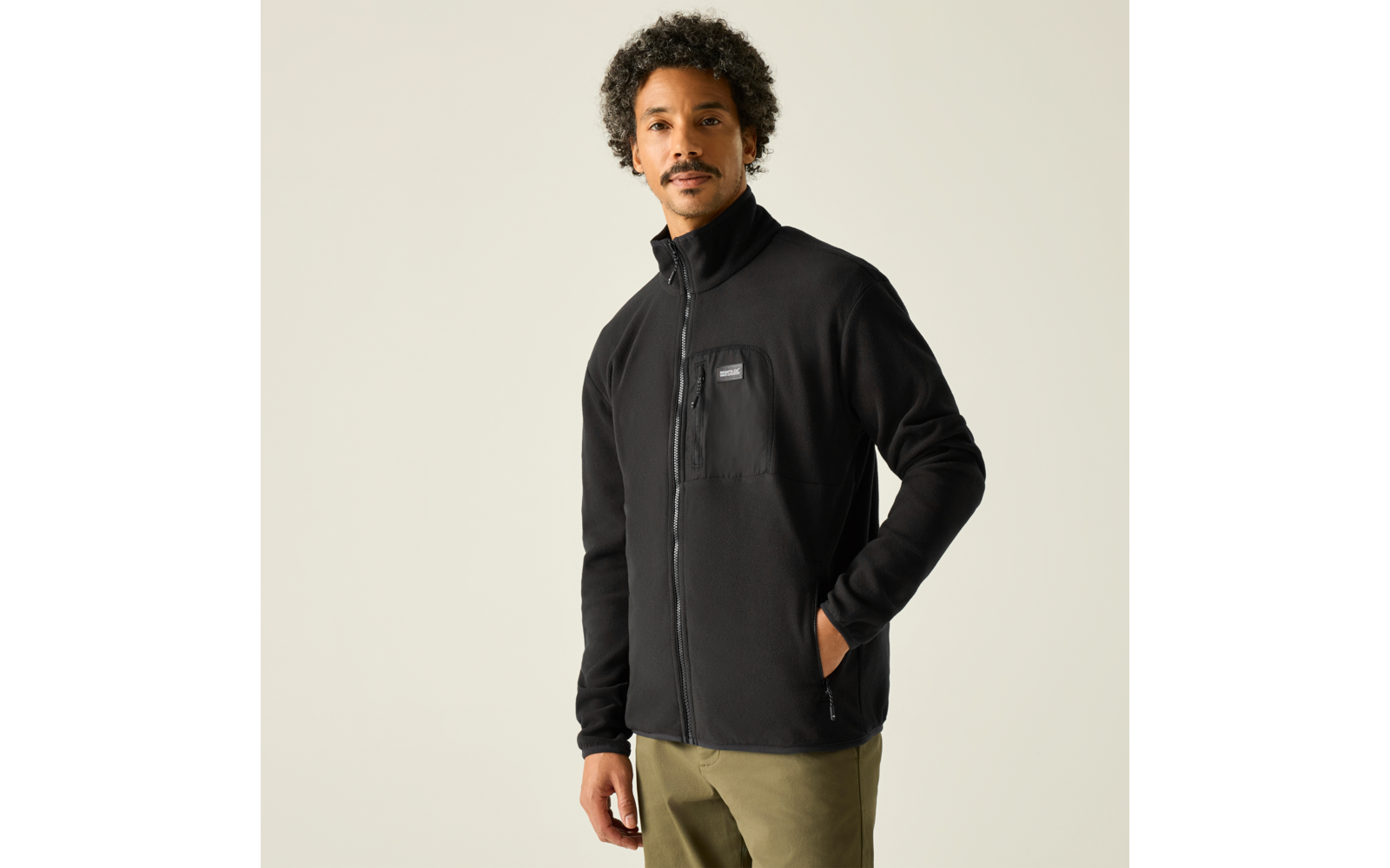 Mann mit lockigem dunklem Haar und leichtem Bart, trägt schwarze Reißverschlussjacke mit Brusttasche und olivgrüne Hose, eine Hand in der Tasche, heller einfarbiger Hintergrund