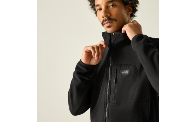 Person mit lockigen Haaren und leichter Gesichtsbehaarung, zieht den Reißverschluss einer schwarzen Fleecejacke mit Brusttasche hoch; Brustbereich vor hellem Hintergrund