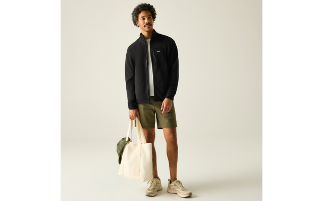 Person steht in schwarzer Jacke mit Reißverschluss, weißem T-Shirt, olivgrünen Shorts und beigen Sneakern, hält eine große beige Tasche mit grüner Kappe vor hellem Hintergrund