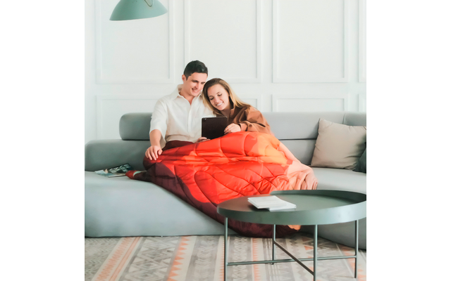 Zwei Personen sitzen zusammen auf einem Sofa, eine Person hält eine Fernbedienung, sie sind mit einer orangefarbenen Decke bedeckt