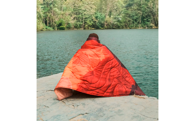 Person sitzt auf einem Felsen am Wasser und ist in eine rote Decke gehüllt