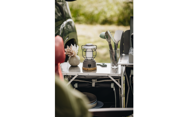 Table pliante avec une lanterne, un verre contenant des ustensiles de cuisine, un petit vase avec des fleurs séchées et une voiture verte en arrière-plan