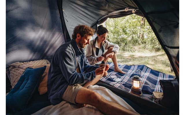 Binnen in een tent zitten twee personen op een geruite deken en houden bekers vast; een lantaarn naast hen; bomen buiten zichtbaar