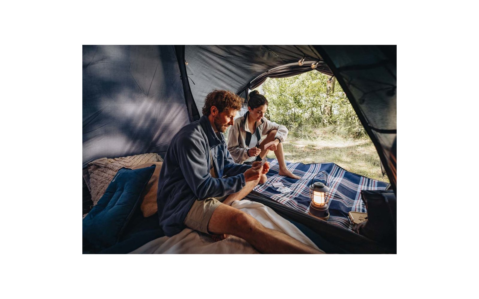 Binnen in een tent zitten twee personen op een geruite deken en houden bekers vast; een lantaarn naast hen; bomen buiten zichtbaar
