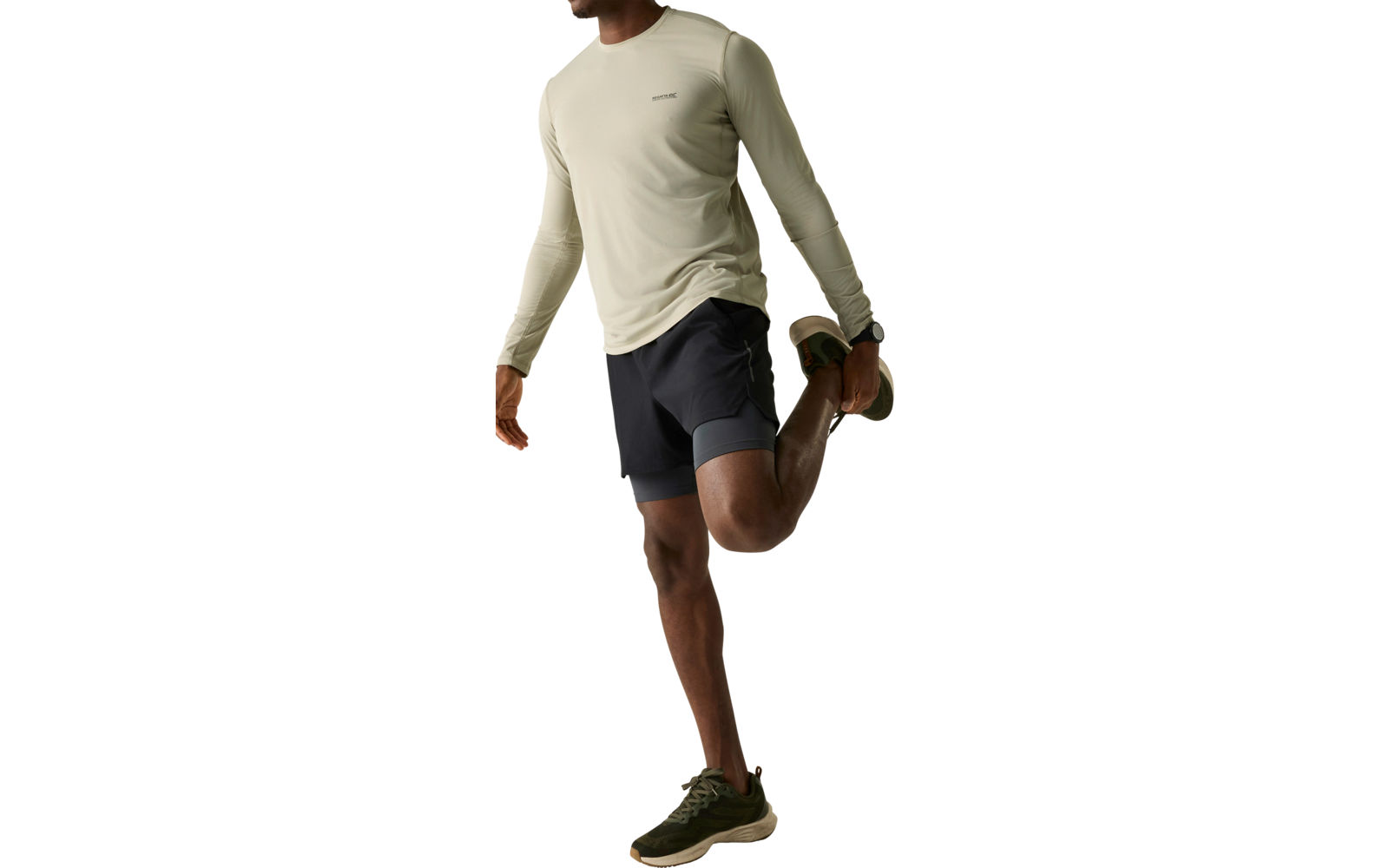 Mann steht auf einem Bein und hält mit einer Hand seinen hinteren Fuß, trägt helles Langarmshirt, dunkle Shorts und grüne Sportschuhe, heller einfarbiger Hintergrund