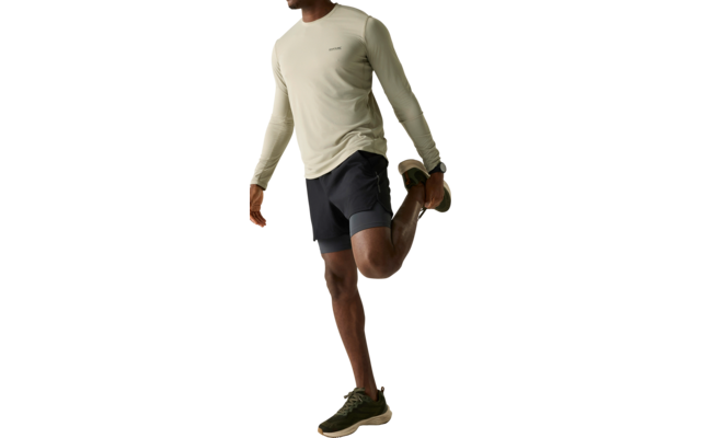 Mann steht auf einem Bein und hält mit einer Hand seinen hinteren Fuß, trägt helles Langarmshirt, dunkle Shorts und grüne Sportschuhe, heller einfarbiger Hintergrund