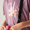 Oberkörper in violettem Shirt mit schwarzem Schultergurt; Aufdruck einer weißen Skelett-Hand mit zwei erhobenen Fingern und Blumen, Schriftzug "DARE2B"; Hände halten ein rosa Objekt