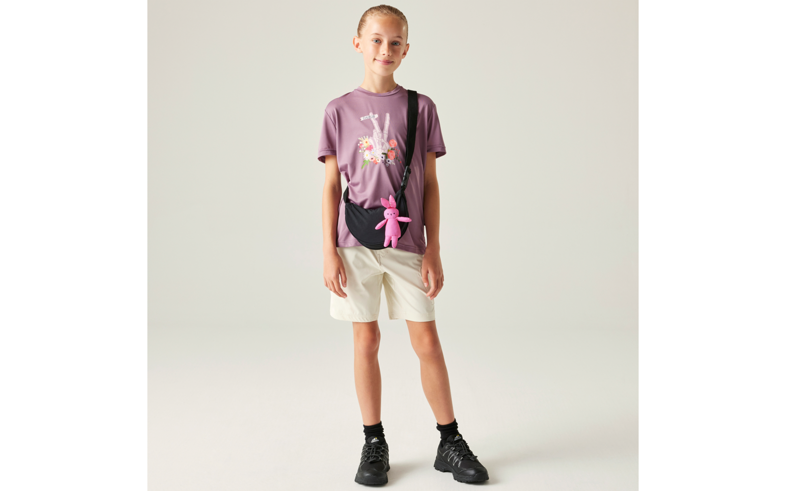 Kind steht vor hellem Hintergrund, trägt lila T‑Shirt mit Blumenmotiv, helle Shorts, schwarze Sneakers und Socken, schwarze Umhängetasche mit rosa Plüschhase