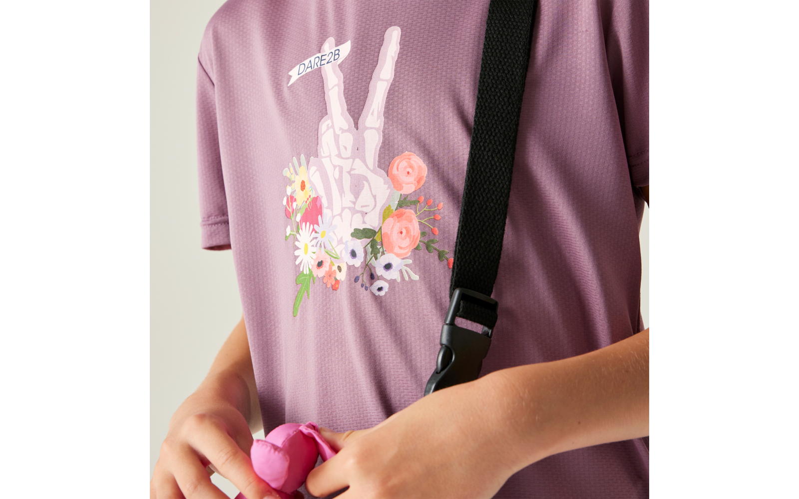 Oberkörper in violettem Shirt mit schwarzem Schultergurt; Aufdruck einer weißen Skelett-Hand mit zwei erhobenen Fingern und Blumen, Schriftzug "DARE2B"; Hände halten ein rosa Objekt
