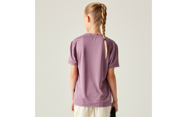 Person von hinten, trägt lila T‑Shirt und helle Hose, Haare zu einem Zopf geflochten, hält ein dunkles Objekt in der rechten Hand, einfarbiger heller Hintergrund