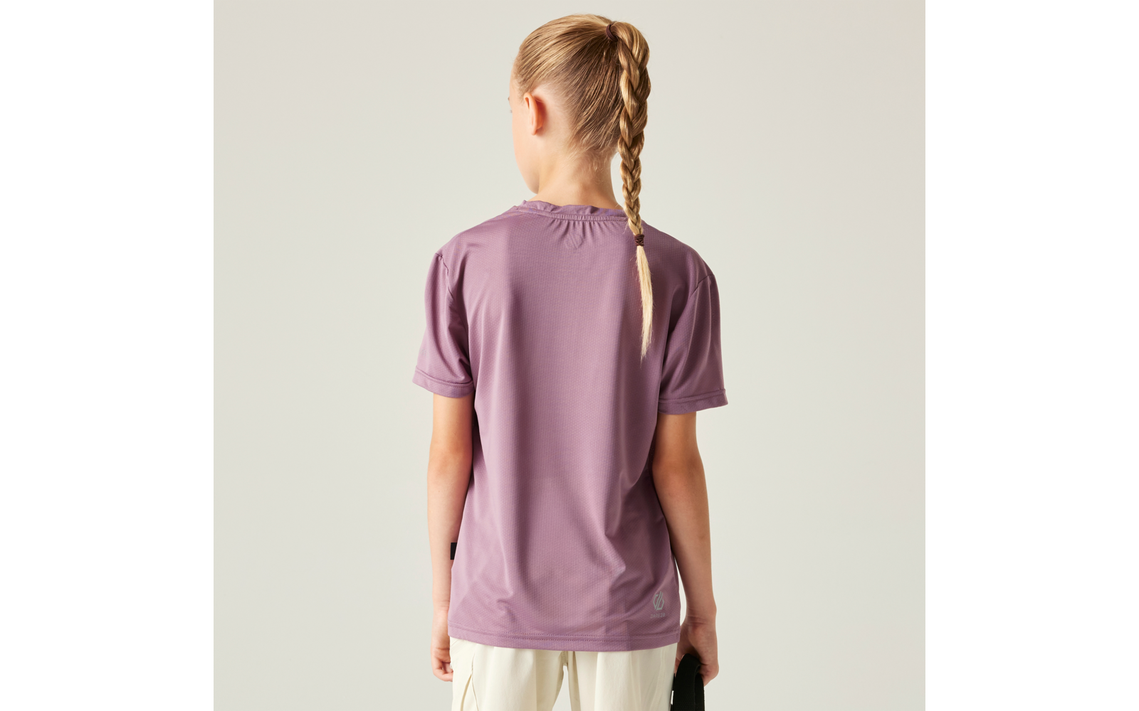 Person von hinten, trägt lila T‑Shirt und helle Hose, Haare zu einem Zopf geflochten, hält ein dunkles Objekt in der rechten Hand, einfarbiger heller Hintergrund