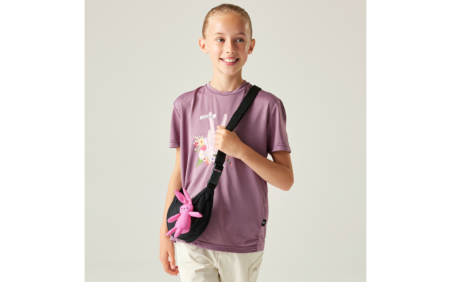 Mädchen mit zurückgebundenem Haar, violettes T‑Shirt mit Motiv, helle Hose, schwarze Umhängetasche mit rosa Stoffhase, steht vor heller einfarbiger Wand und hält den Taschengurt